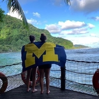 two girls facing the water with large flag of M draped across shoulders